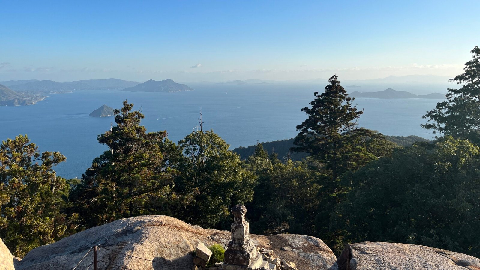 Standing at the top of Mount Misen, overlooking conifer trees descending a mountainside until Miyajima Island ends. Ocean then expands, with other small islands dotted along until the photo is swallowed under the horizon.